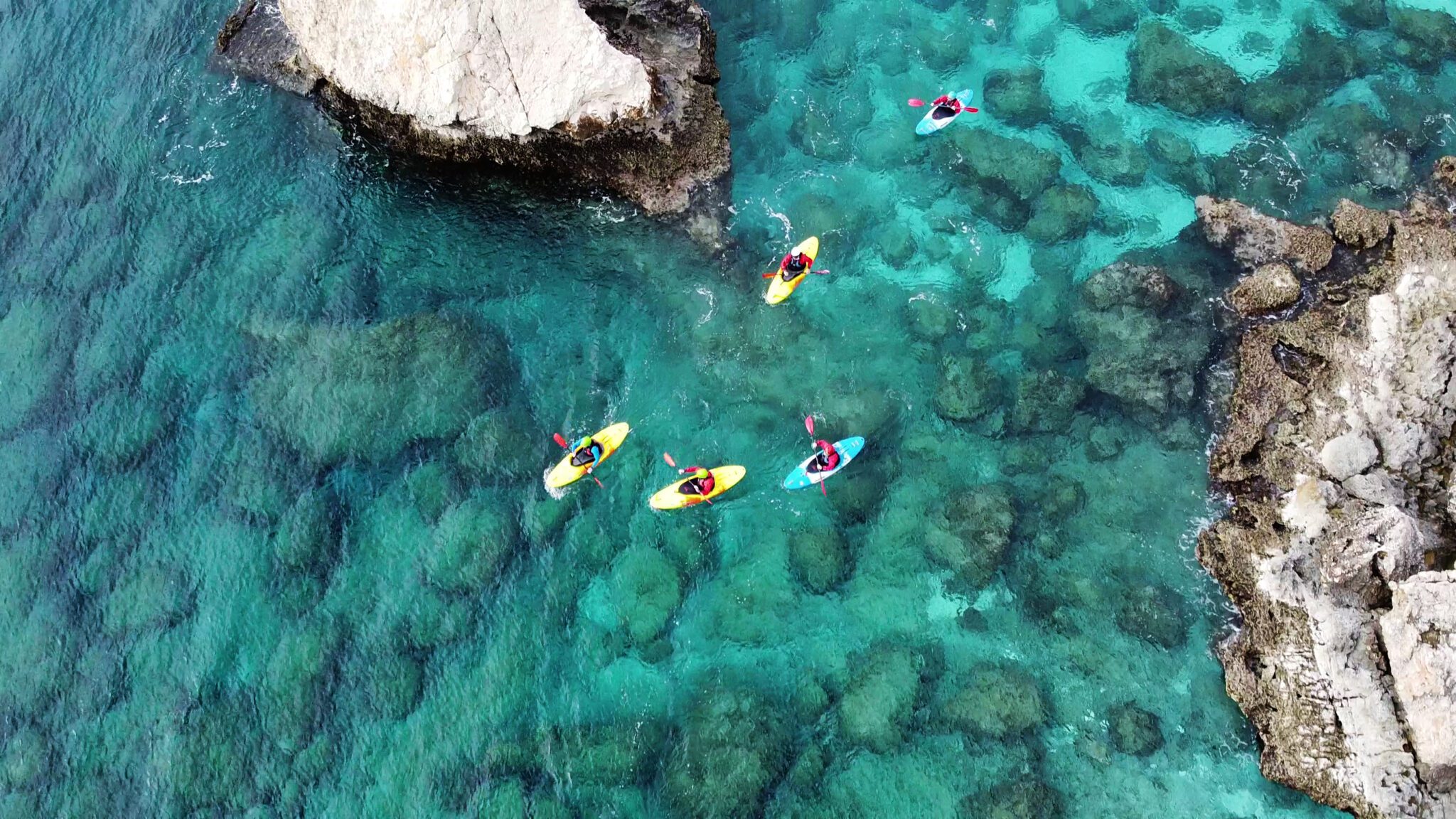 Kayakers paddling in clear turquoise water near a rocky shore.