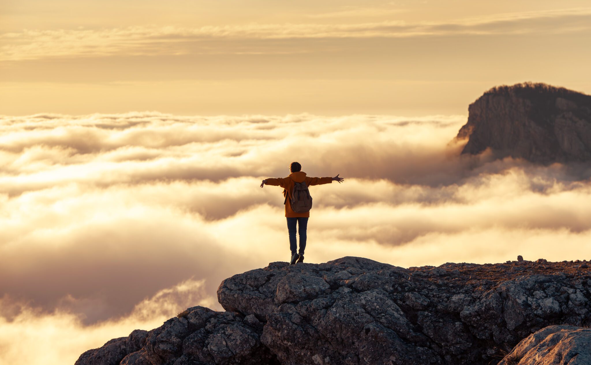 Person standing on a cliff above the clouds at sunrise.