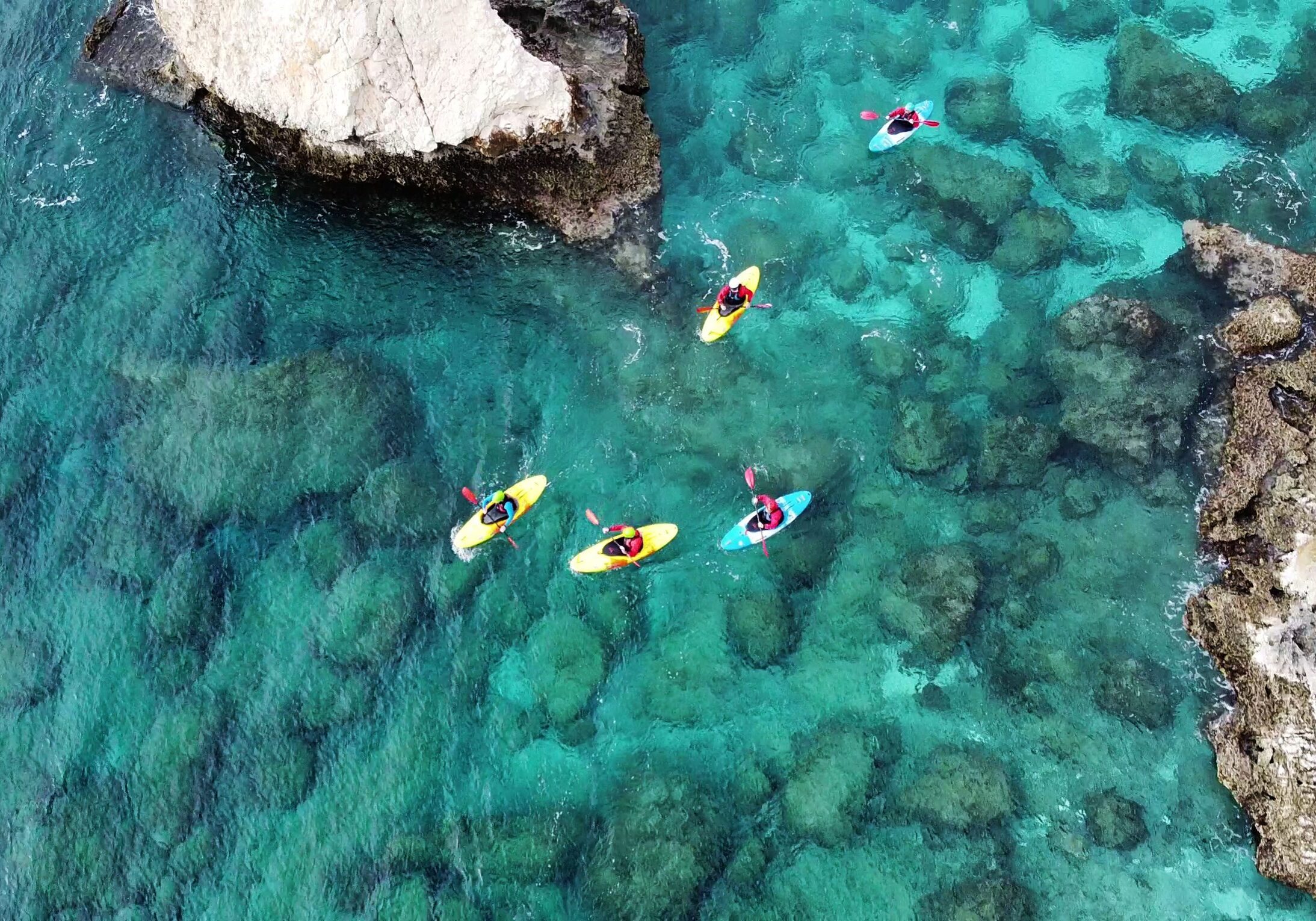 Kayakers paddling in clear turquoise water near a rocky shore.