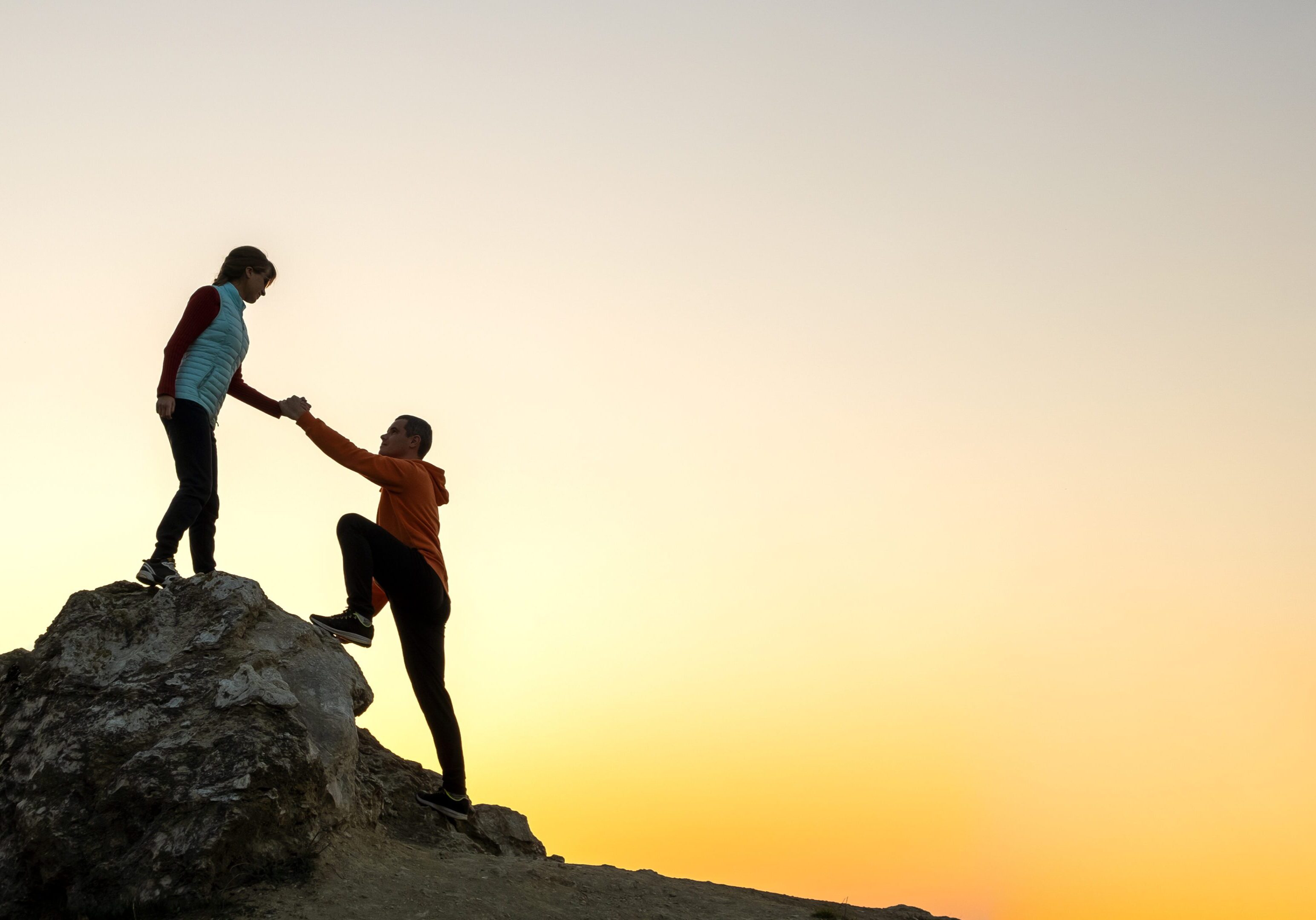 One person helps another climb a rocky hill at sunset.